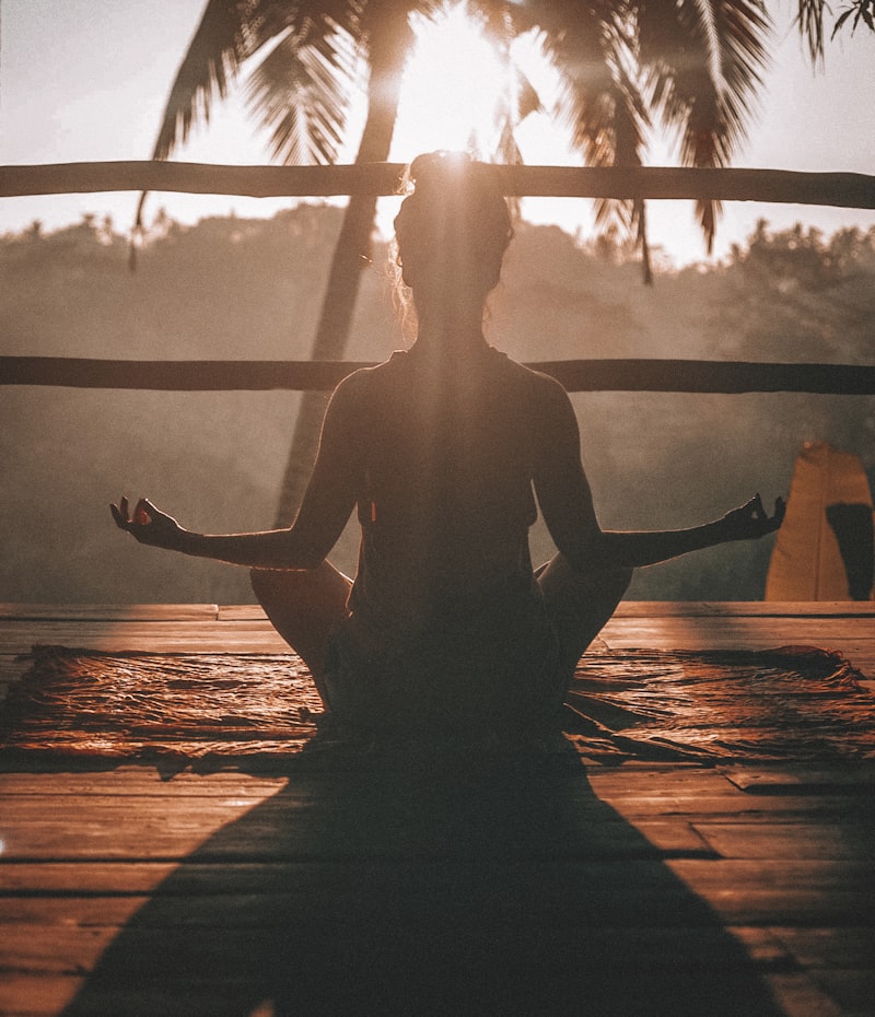 Silhouette of a person meditating in cross-legged pose on a wooden deck at golden hour with palm trees