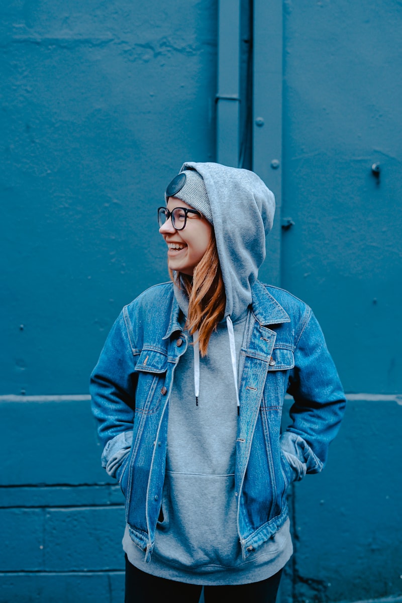 Smiling young woman in a denim jacket and grey hooded sweatshirt standing against a blue wall